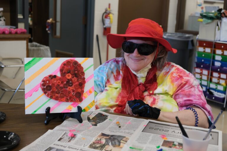 resident dressed in tie dye and vibrant reds holding up their artwork that is as colorful as themselves with a big heart in the middle
